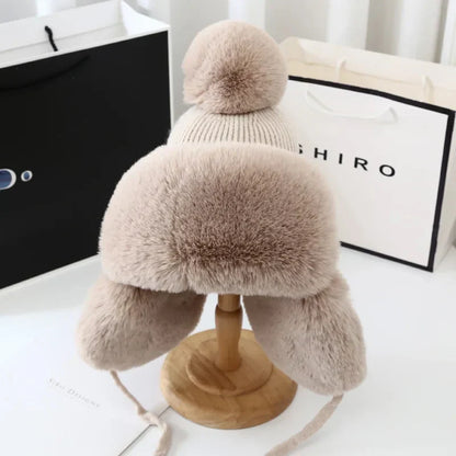 Beige Wintermütze mit Kunstfell, Bommel und Ohrenklappen auf Holzständer, modisch und warm.