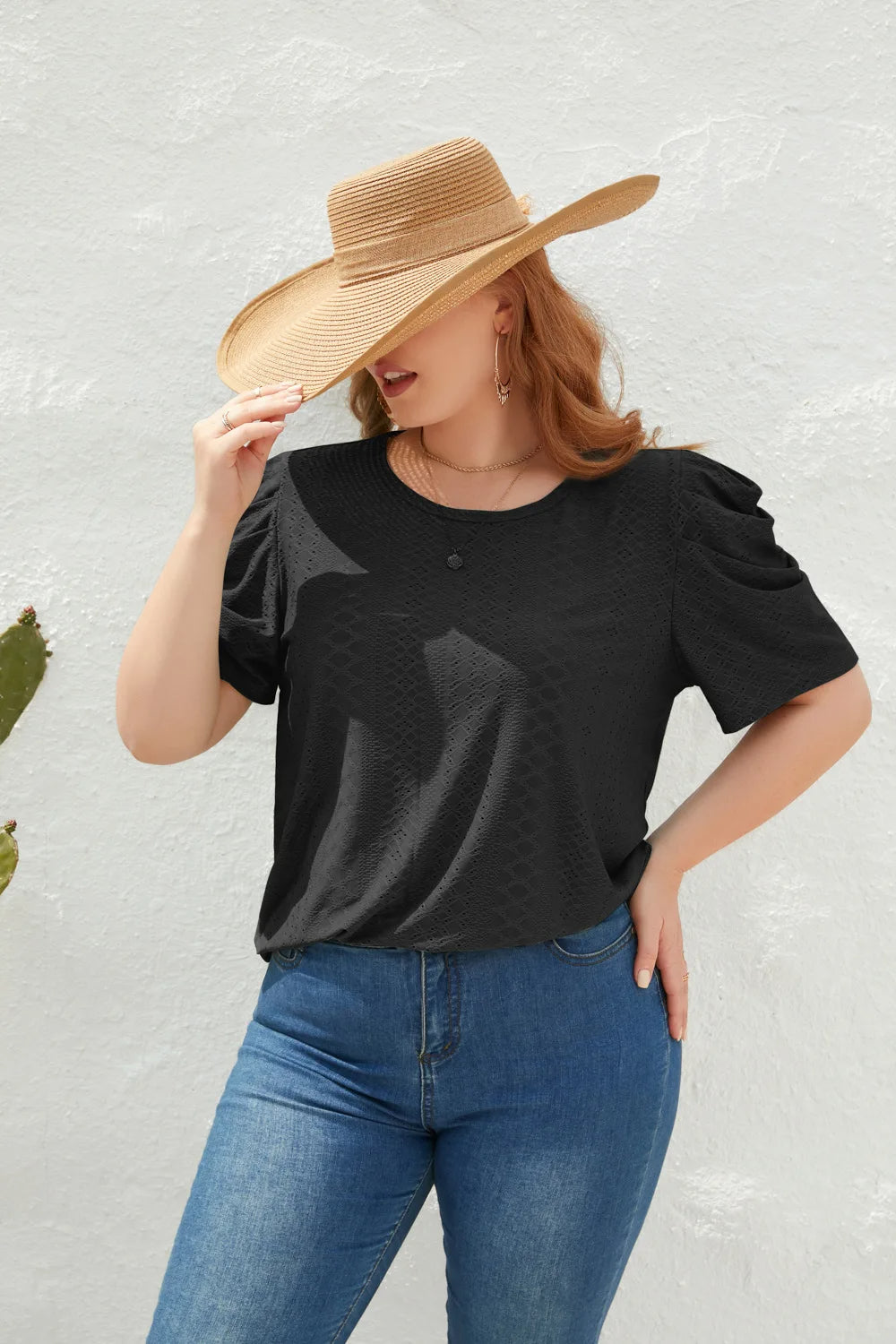 Frau mit schwarzem T-Shirt, blauer Jeans und Strohhut vor weißer Wand.