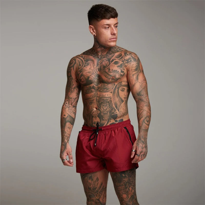 Mann mit Tattoos trägt rote Sportshorts, muskulös, Fitnessmode, Studioaufnahme.