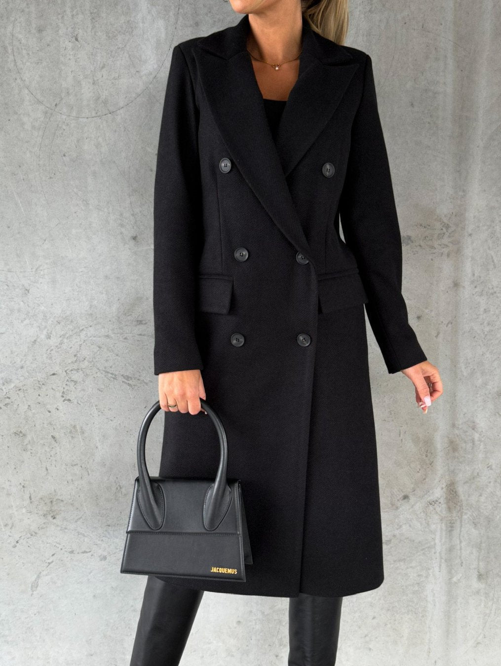 Frau in schwarzem Wollmantel und Lederstiefeln mit schwarzer Handtasche vor grauer Wand.
