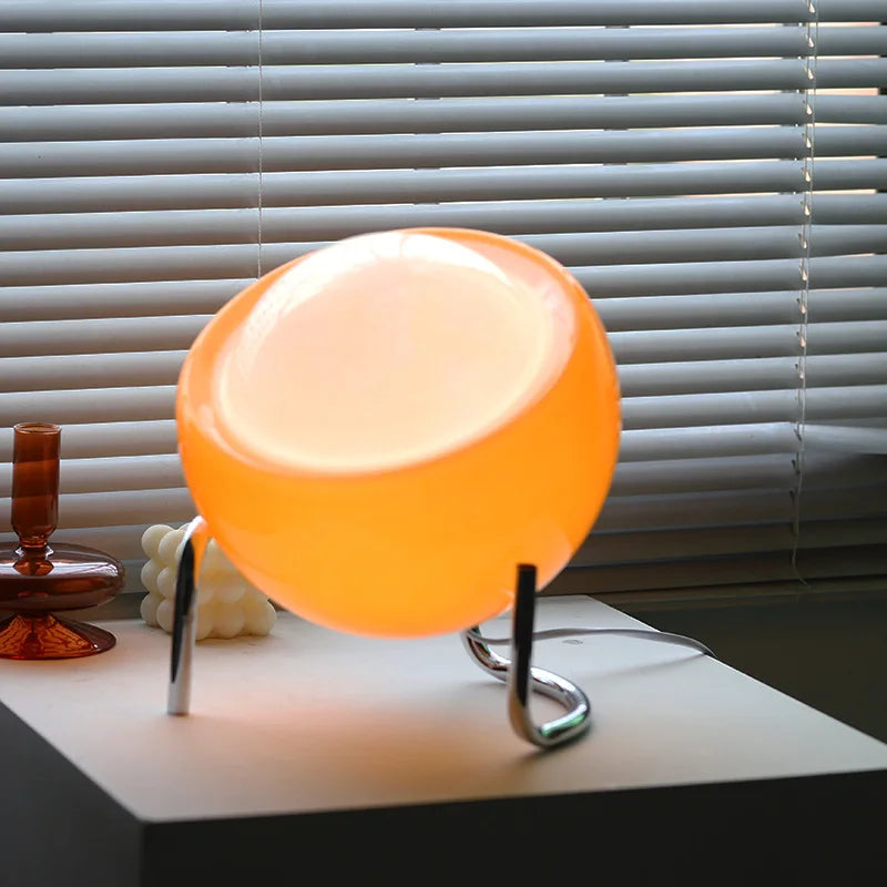 Orange Tischlampe aus Glas auf weißem Tisch, modernes Design, vor Jalousien im Sonnenlicht.
