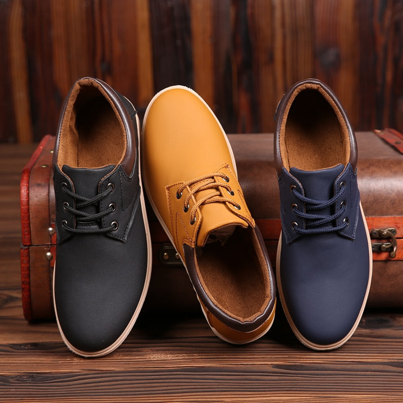 Drei Paar Herren-Ledersneaker in Schwarz, Gelb und Blau auf Holzhintergrund vor Koffer.