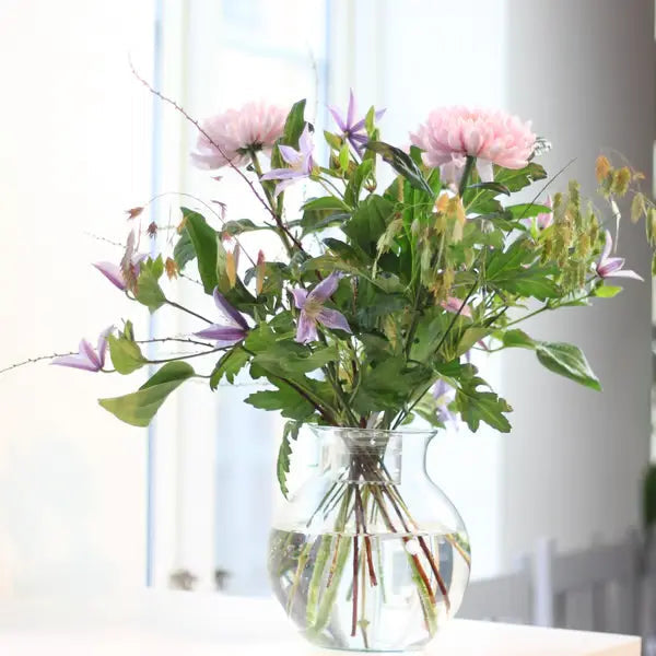 Bukett med rosa blommor, lila klematis och gröna blad i en rund glasvas med vatten vid ett fönster. 4