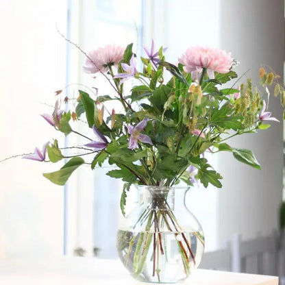 Bukett med rosa blommor, lila klematis och gröna blad i en rund glasvas med vatten vid ett fönster. 4