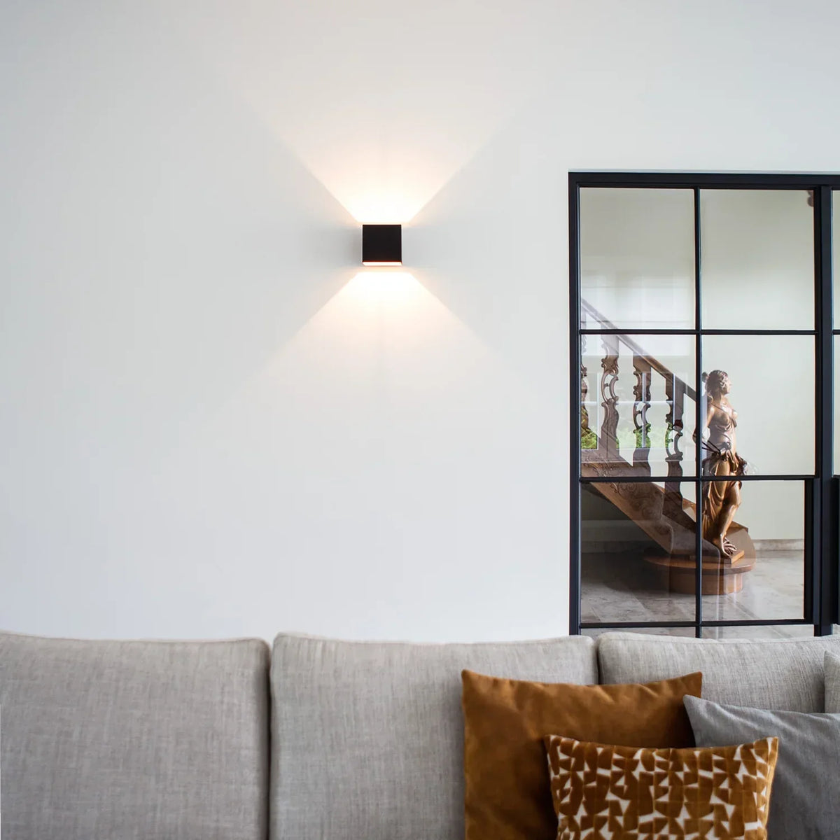Svart vägglampa på en vit vägg ovanför en grå soffa med orange kuddar, bredvid en svartinramad glasdörr. 10