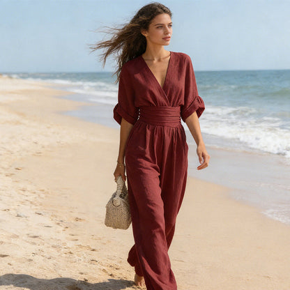 Kvinna i vinröd jumpsuit promenerar barfota på en strand med flätad väska, håret blåser i vinden. 7