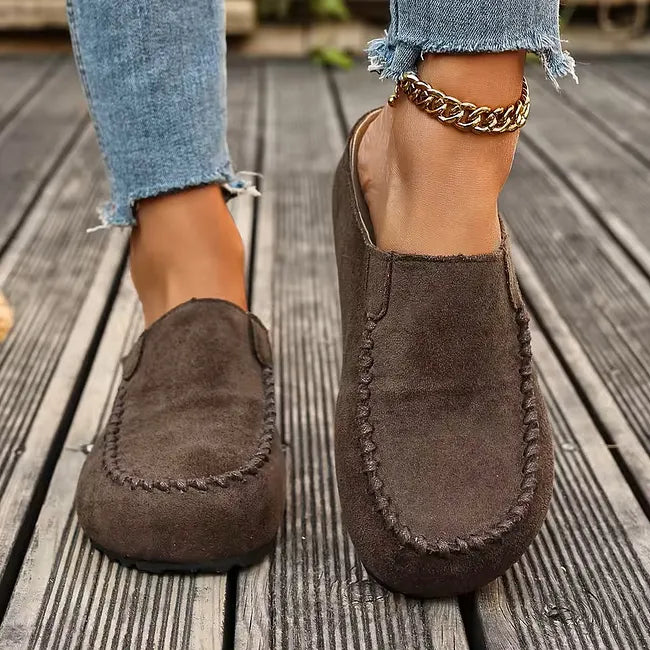 Framifrån: mörkbruna tofflor med sömdetaljer vid tån, bärs av en modell i fransiga jeans och guldfärgat ankelkedja på träbrygga. 4