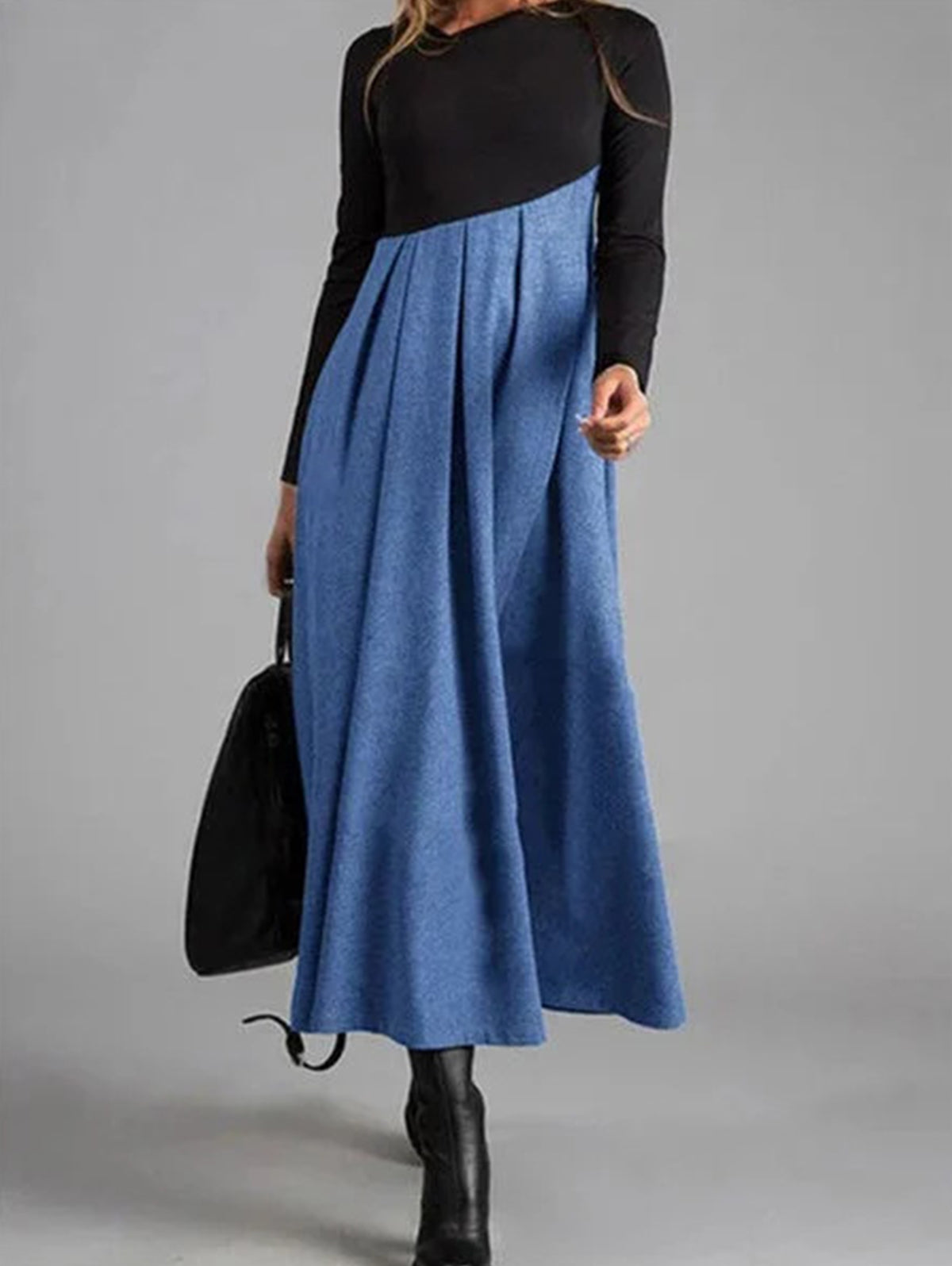 Frau in langem, blauem Kleid mit schwarzem Oberteil, hält schwarze Tasche, trägt schwarze Stiefel.