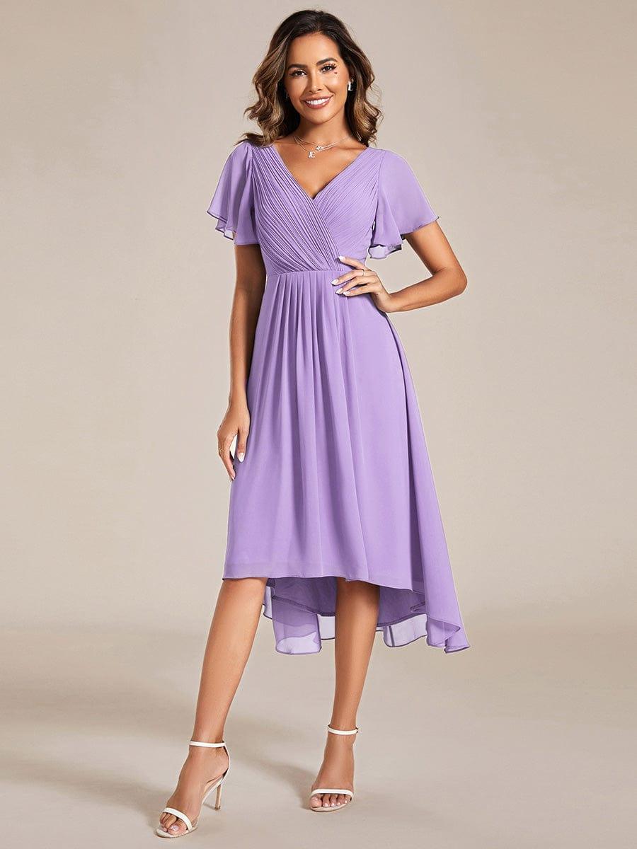 Lila klänning med draperad V-ringning, korta volangärmar och asymmetrisk fåll, modellen ler med handen på höften, framifrån. 100