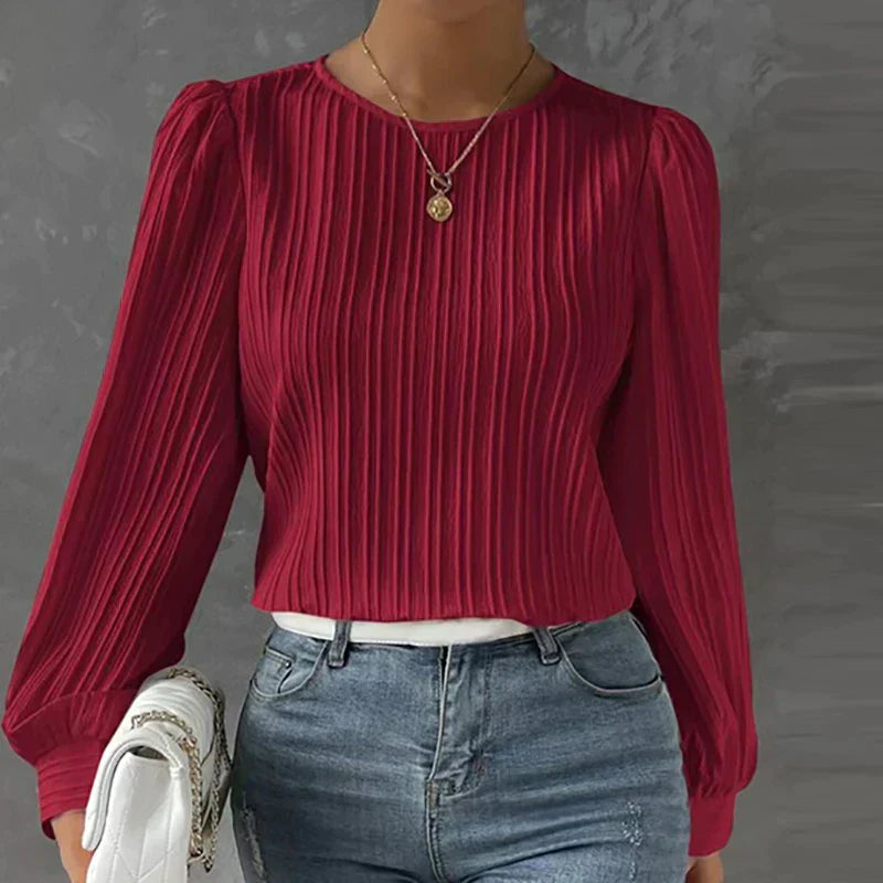Frau in roter, plissierter Bluse mit langen Ärmeln und blauer Jeans vor grauem Hintergrund.