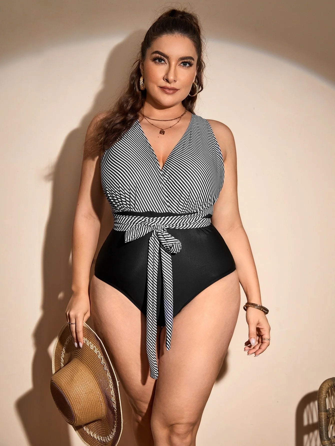 Frau im gestreiften Badeanzug mit Gürtel, hält Strohhut, Plus-Size-Mode, Sommerkleidung.