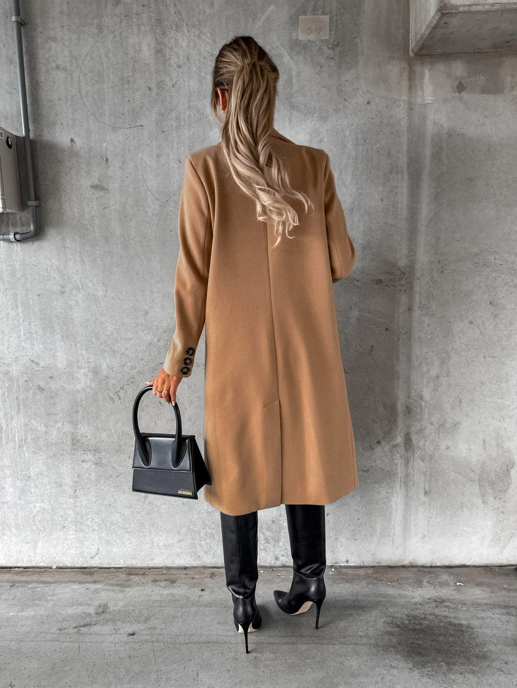Frau in beigem Mantel, schwarze Stiefel, elegante Handtasche, Betonhintergrund.