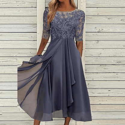 Frau in elegantem, blauem Sommerkleid mit Spitze, knielang, vor Holzwand.