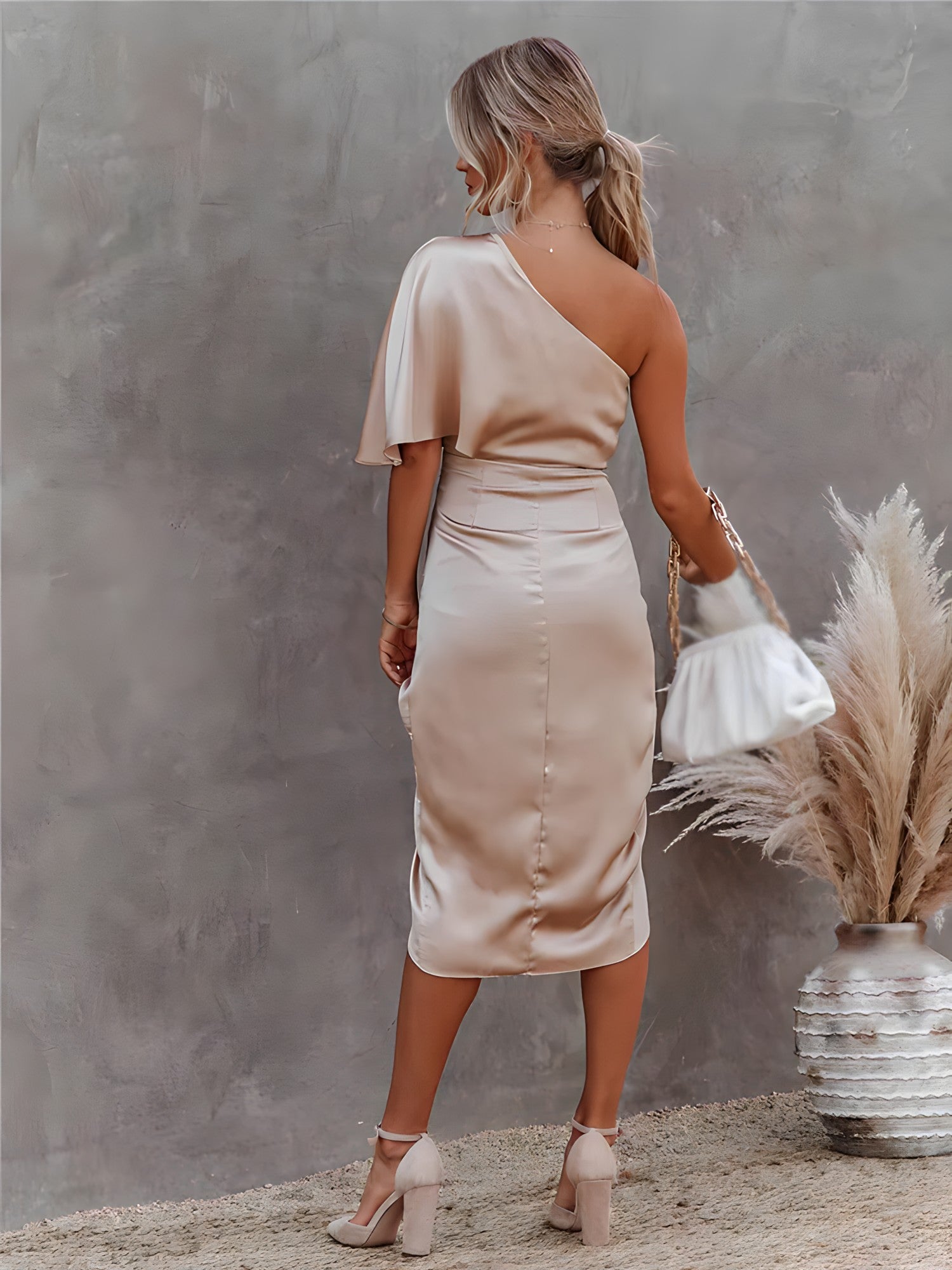 Frau in eleganter, einseitiger, beiger Satin-Kleid, hält weiße Handtasche, steht vor grauer Wand.