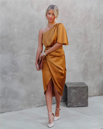 Frau in einem goldenen, schulterfreien Seidenkleid mit Clutch und weißen High Heels vor grauer Wand.