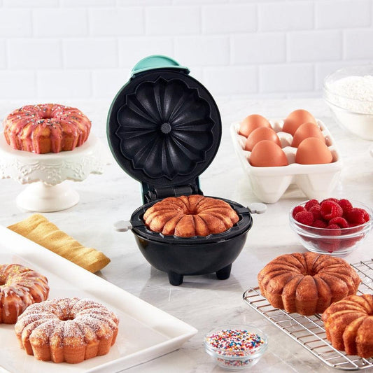 Elektrische Kuchenbackmaschine, schwarz, mit gebackenem Kuchen, umgeben von Eiern und Zutaten.