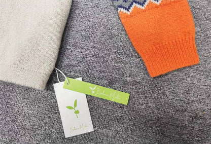 Zwei Pullover auf grauem Teppich, einer beige, einer orange mit Zickzackmuster, Etikett sichtbar.