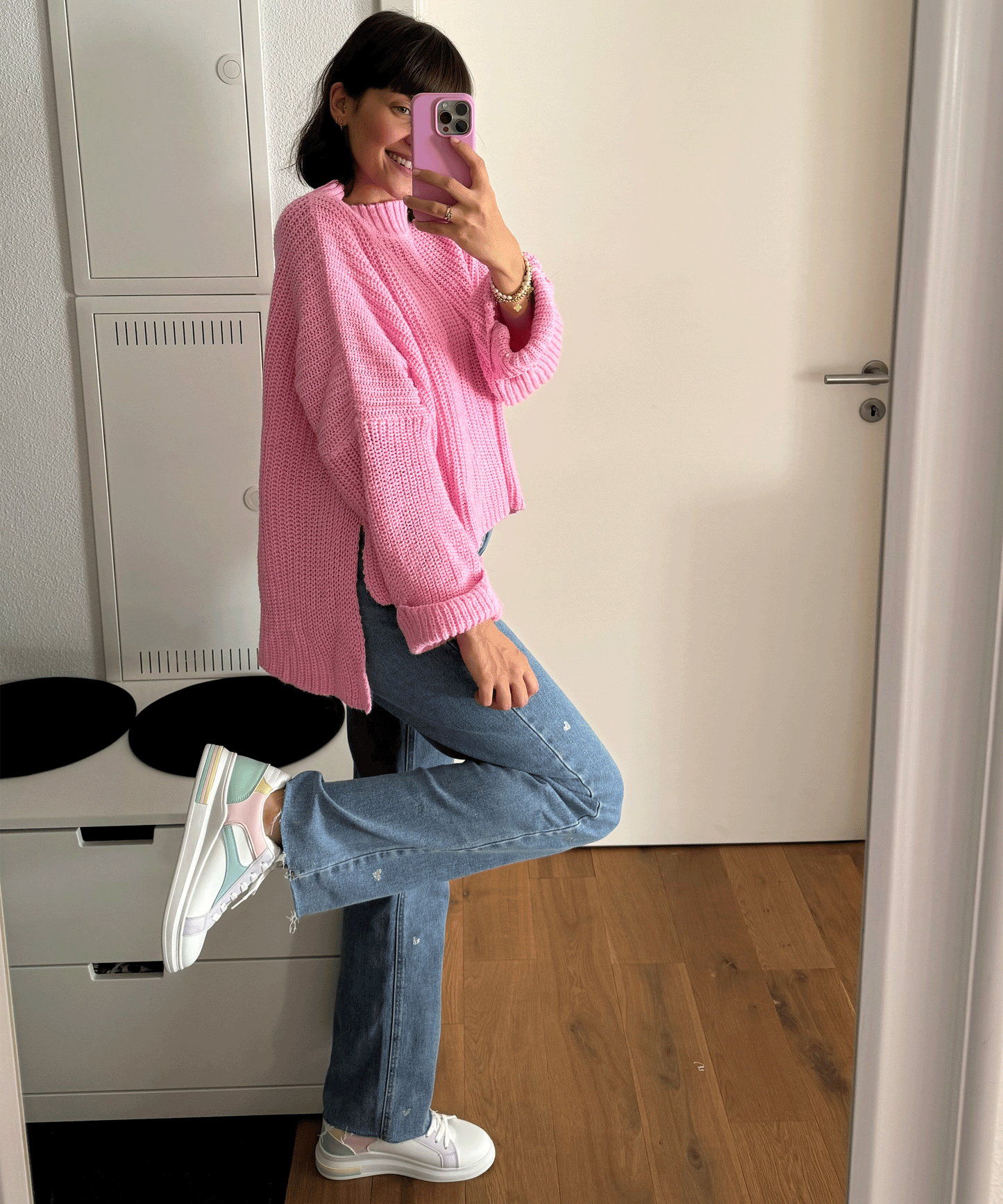 Frau in rosa Pullover und blauen Jeans, trägt weiße Sneakers, macht Selfie im Spiegel.