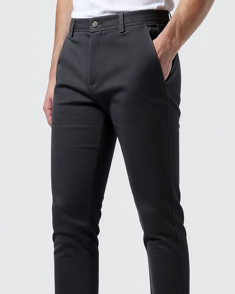 Närbild av mörkgrå chinos snett framifrån med knapp i midjan och handen i sidfickan, modell i vit t-shirt. 10