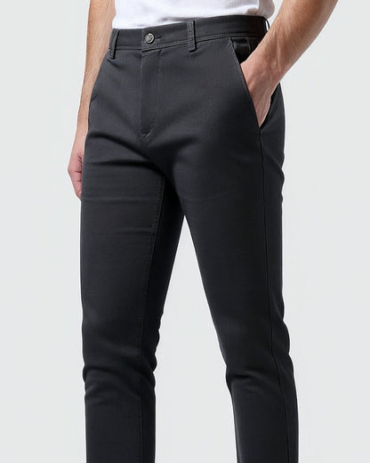 Närbild av mörkgrå chinos snett framifrån med knapp i midjan och handen i sidfickan, modell i vit t-shirt. 10