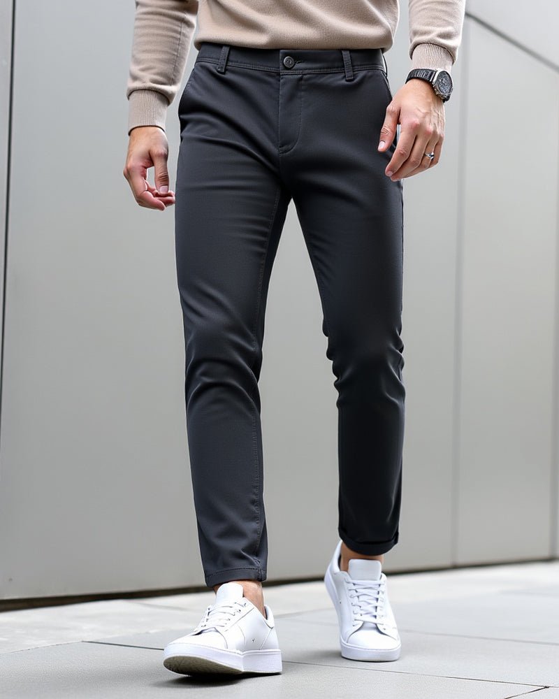 Mörkgrå chinos med beige stickad tröja och vita sneakers, utomhus mot en grå vägg med uppvikta byxben. 9