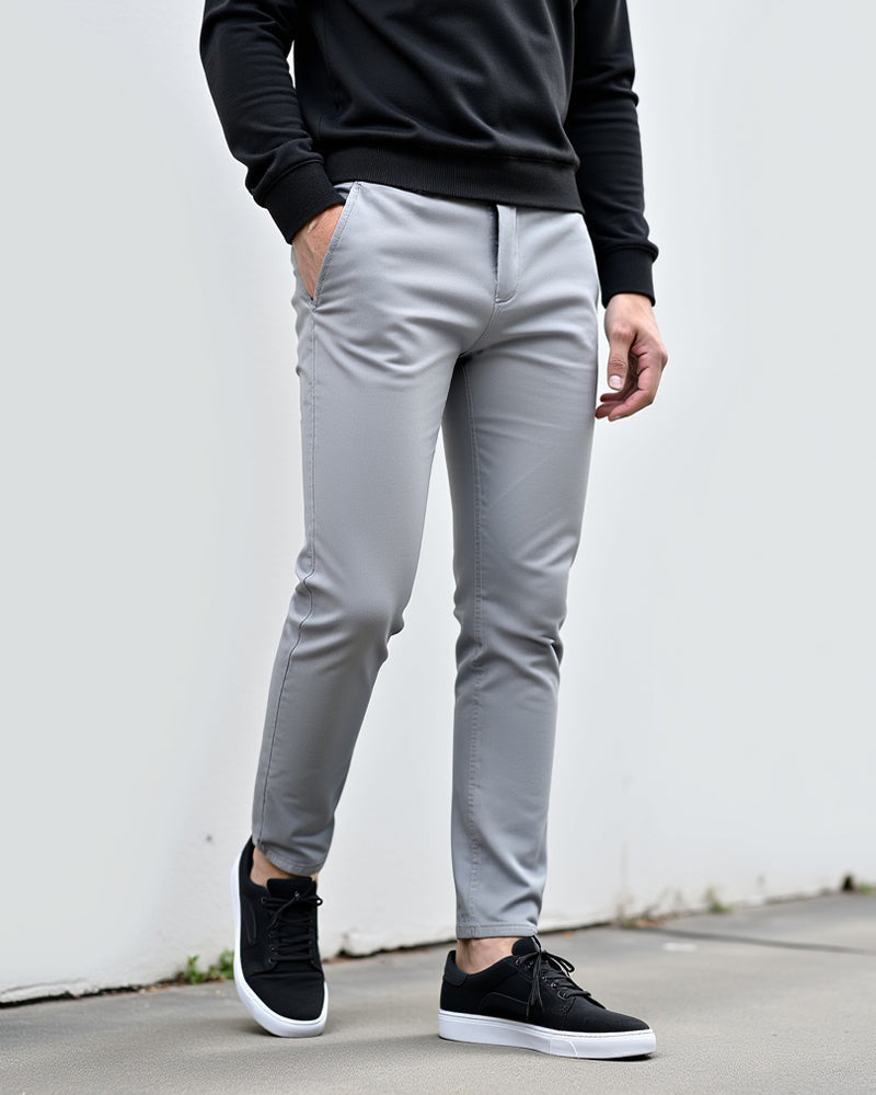 Ljusgrå chinos med svart tröja och svarta sneakers, stylade utomhus mot en vit vägg med handen i fickan. 2