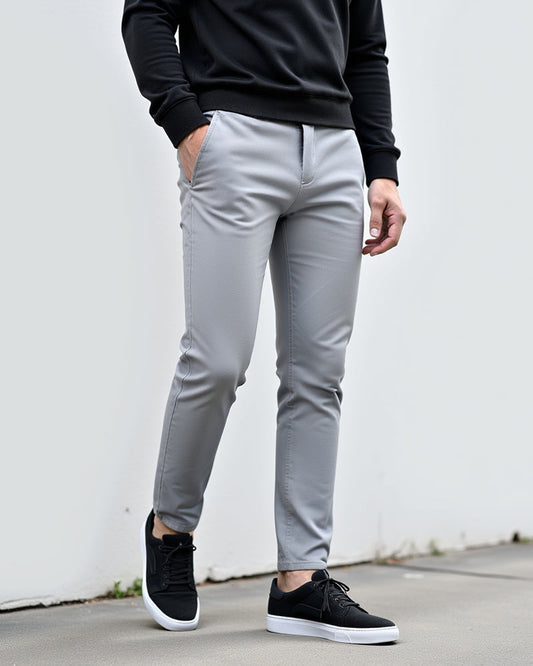 Ljusgrå chinos med svart tröja och svarta sneakers, stylade utomhus mot en vit vägg med handen i fickan. 2