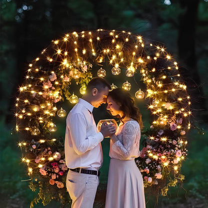 Paar unter beleuchtetem Blumenbogen, romantische Hochzeit, Abend, Lichter, Naturkulisse.