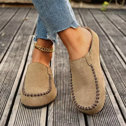 Snett framifrån: beige tofflor med bruna sömmar och platt sula, bärs av en modell i fransiga jeans på träbrygga. 5