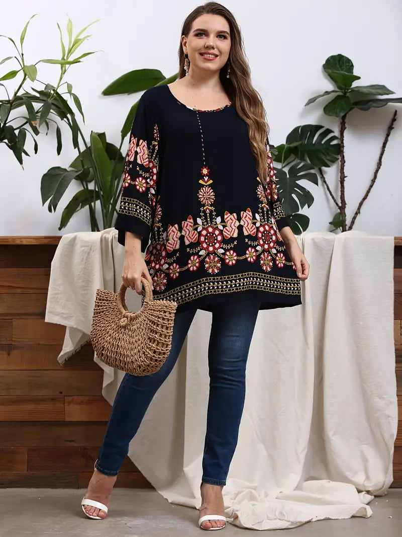 Frau in schwarzer Tunika mit Blumenmuster, Jeans und Strohtasche, vor Pflanzen stehend.