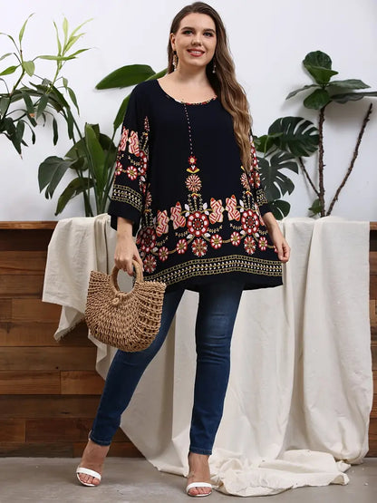 Frau in schwarzer Tunika mit Blumenmuster, Jeans und Strohtasche, vor Pflanzen stehend.