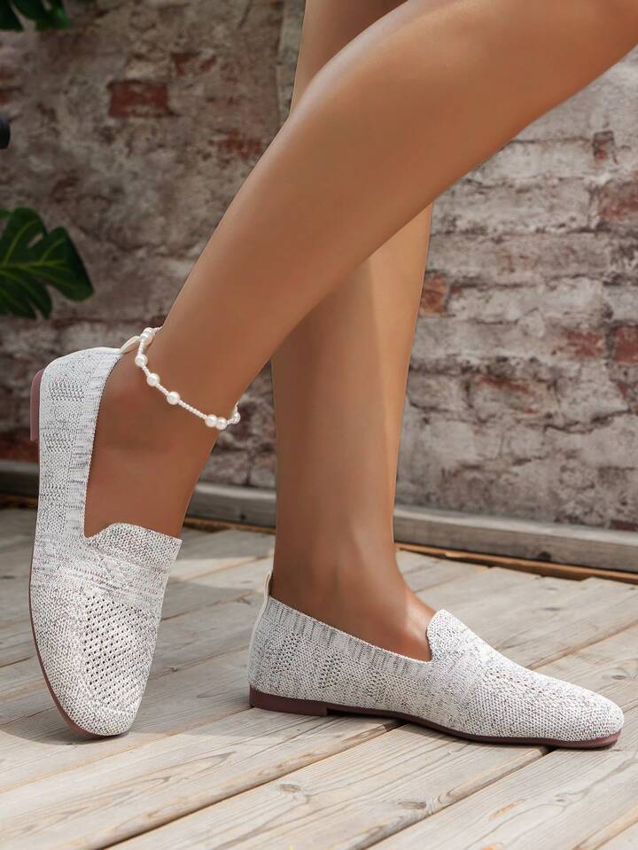 Vitgrå stickade slip-on-skor sedda från sidan på en modell med pärlankelkedja, mot en tegelvägg på trädäck. 14