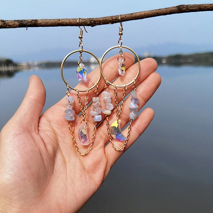 Hand hält goldene Ohrringe mit Kristallen und Ketten vor Seehintergrund. Boho-Schmuck, Damenmode.