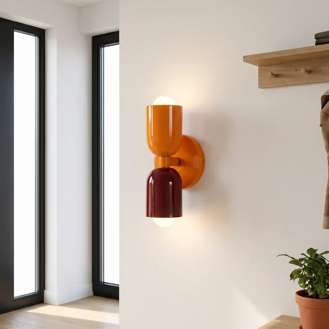 Tänd vägglampa i orange och vinrött i en hall med höga fönster, hylla med krokar och en krukväxt. 11