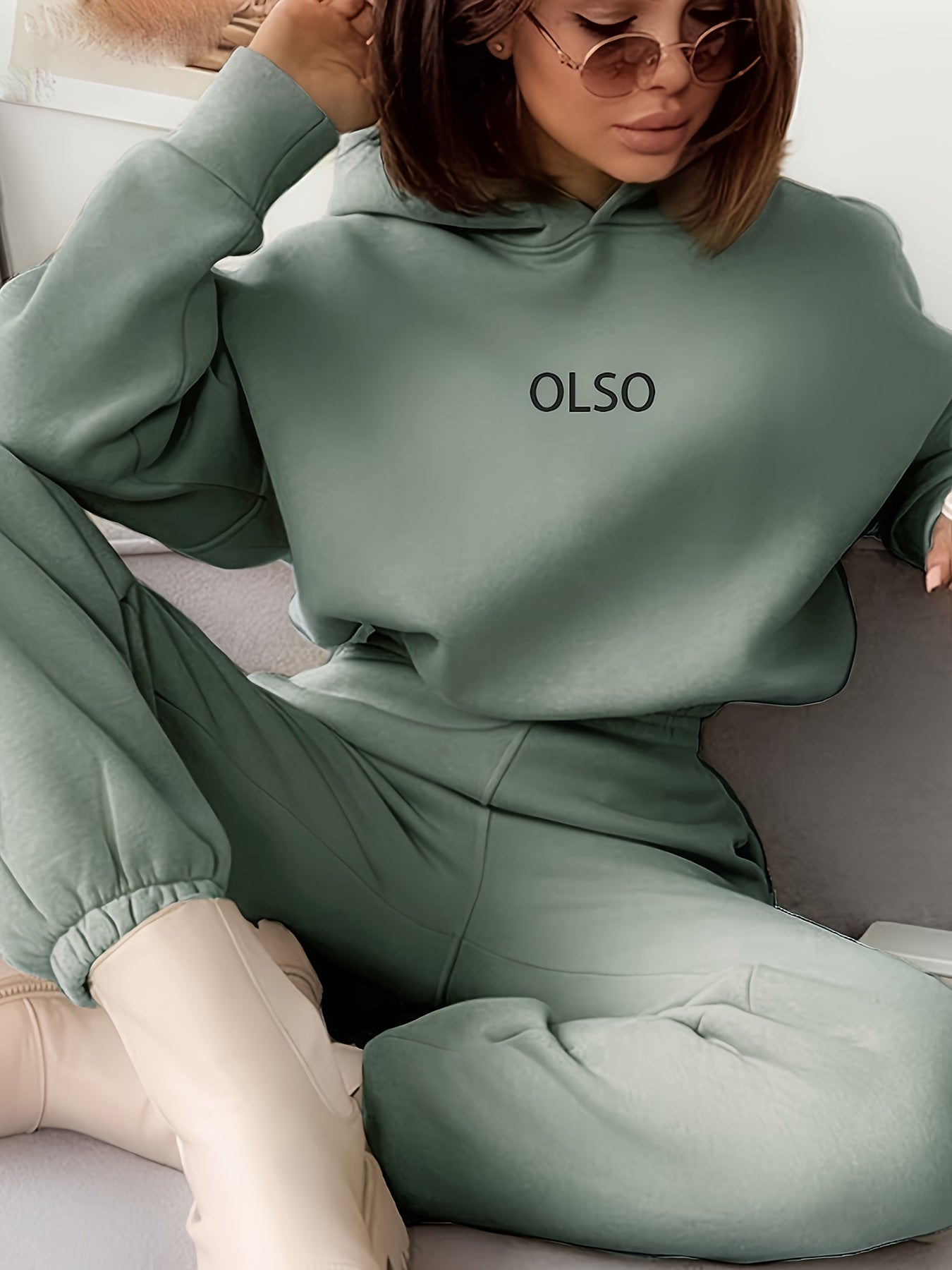 Frau in grünem OLSO-Hoodie und Jogginghose, trägt Sonnenbrille, sitzt auf Sofa.