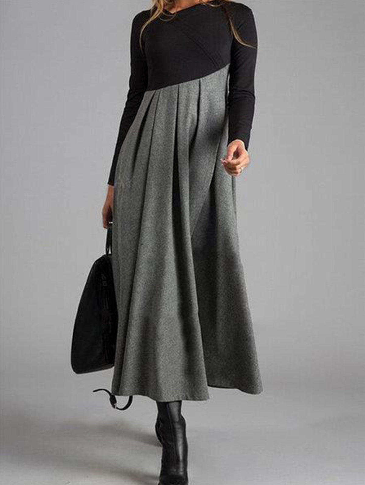 Frau in langem, grauem Maxikleid mit schwarzen Ärmeln und Stiefeln, hält schwarze Tasche.