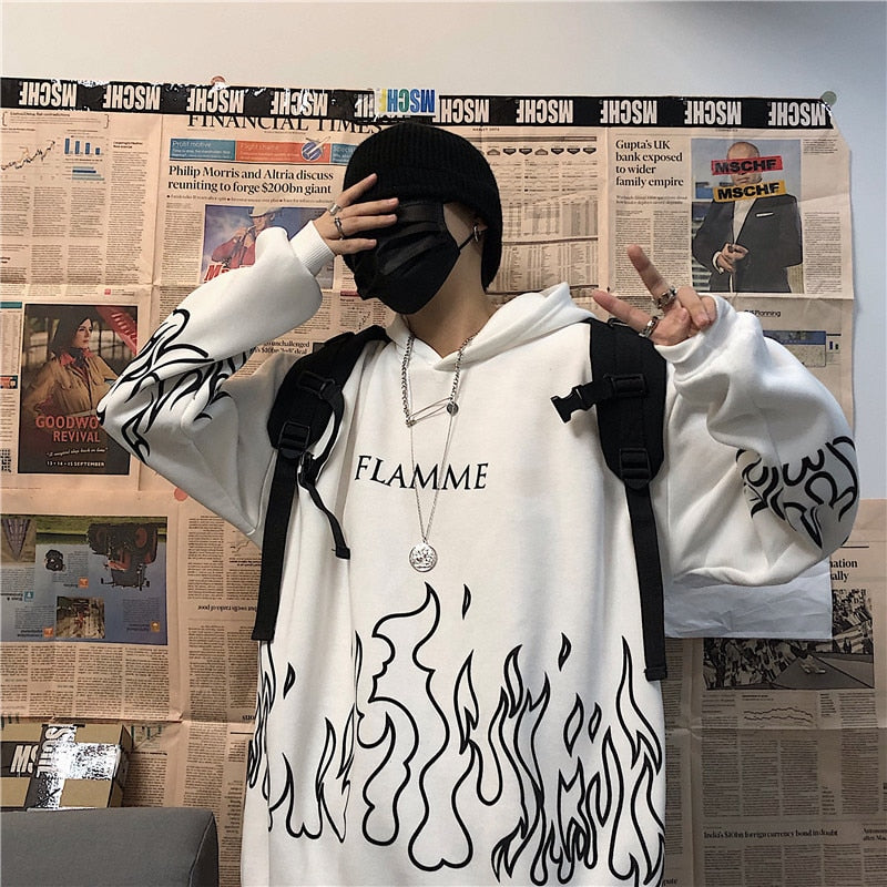 Person in weißem Hoodie mit Flammenmuster, schwarzer Mütze und Maske, vor Zeitungswand.