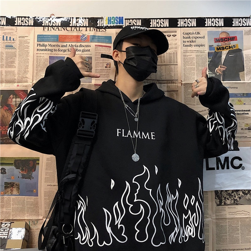 Person in schwarzem Hoodie mit Flammenmuster, schwarzer Maske und Kappe vor Zeitungswand.