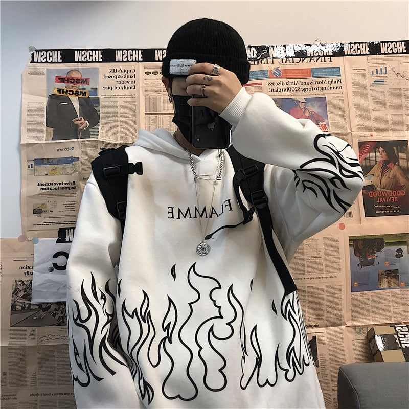 Person in weißem Hoodie mit Flammenmuster, schwarzer Mütze und Rucksack, vor Zeitungswand.