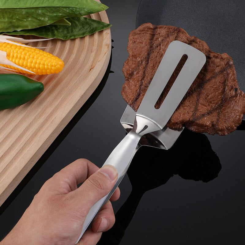Hand hält Edelstahl-Grillwender mit Steak, daneben Maiskolben und grüne Paprika auf Holzbrett.