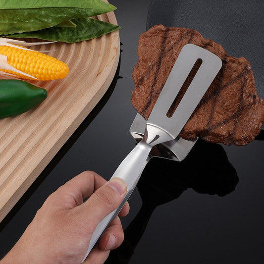 Hand hält Edelstahl-Grillwender mit Steak, daneben Maiskolben und grüne Paprika auf Holzbrett.