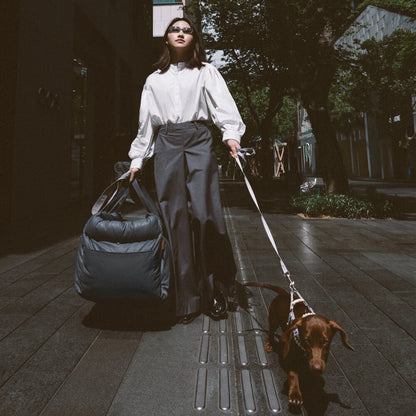 Frau mit Hund und Reisetasche auf Gehweg, modisch gekleidet, Stadtumgebung, Tageslicht.