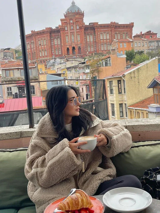 Frau in beiger Teddyjacke trinkt Kaffee auf Terrasse mit Blick auf rotes Backsteingebäude in Istanbul.