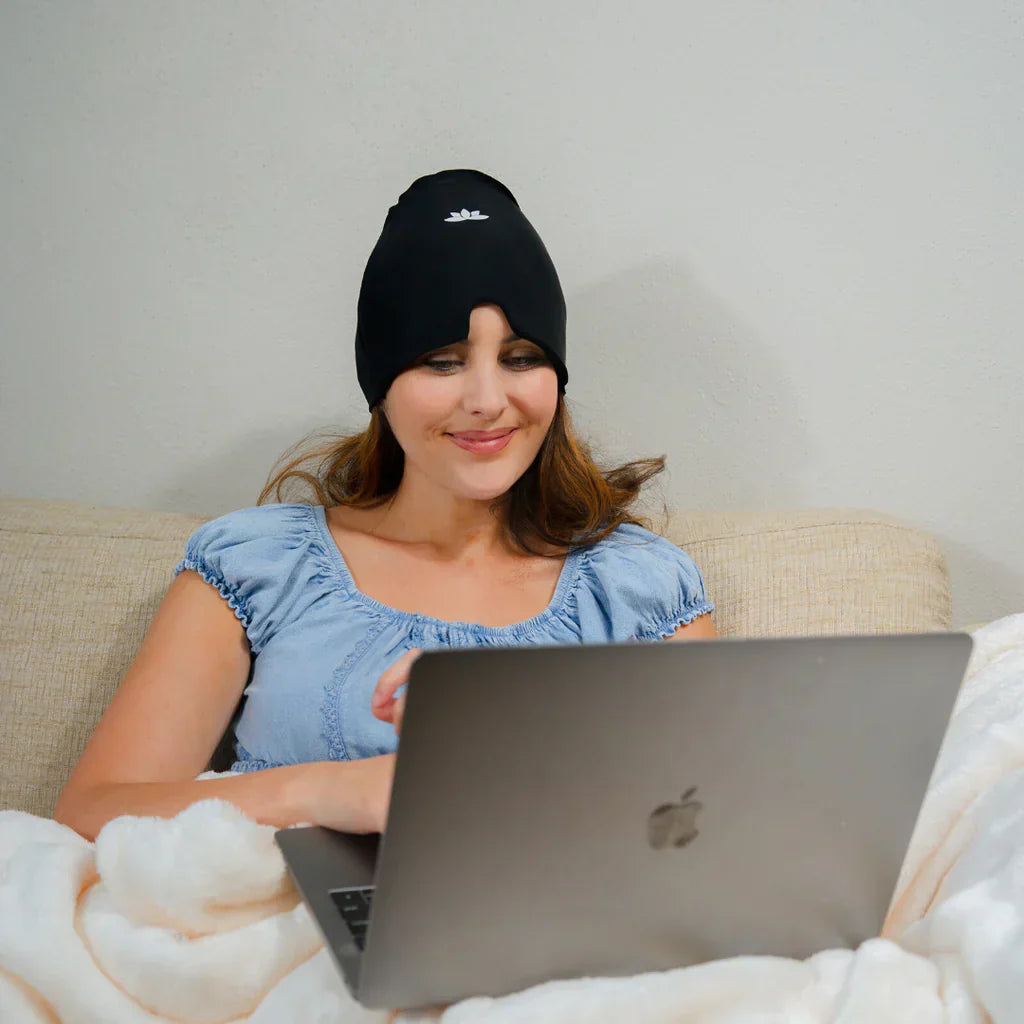 Frau mit schwarzer Kühlmütze arbeitet am Laptop auf Sofa, blaue Bluse, weiße Decke.