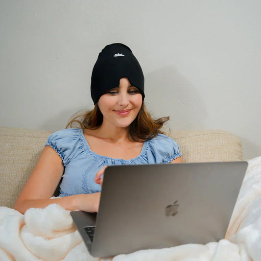 Frau mit schwarzer Kühlmütze arbeitet am Laptop auf Sofa, blaue Bluse, weiße Decke.
