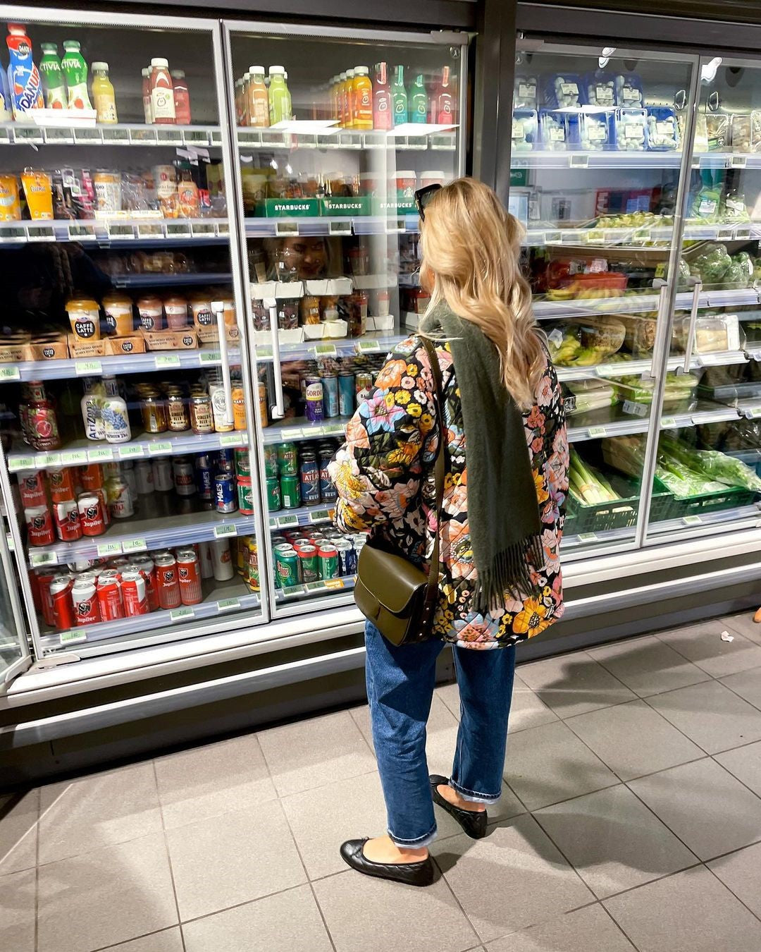 Frau im Supermarkt vor Kühlregal, trägt bunte Jacke, Jeans, schwarze Schuhe, grüne Tasche.