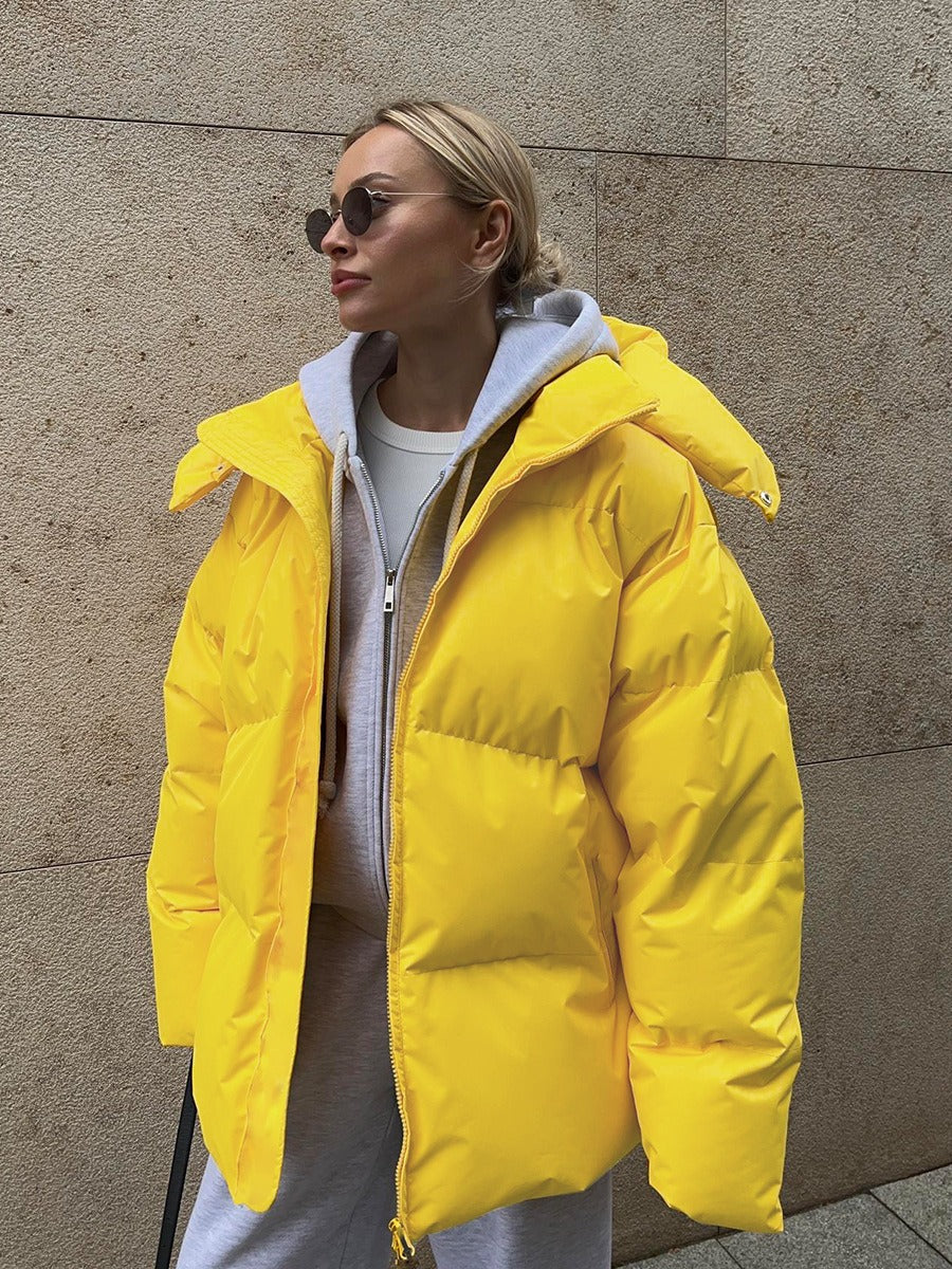 Frau in gelber Daunenjacke, grauem Hoodie und Sonnenbrille vor beiger Wand.