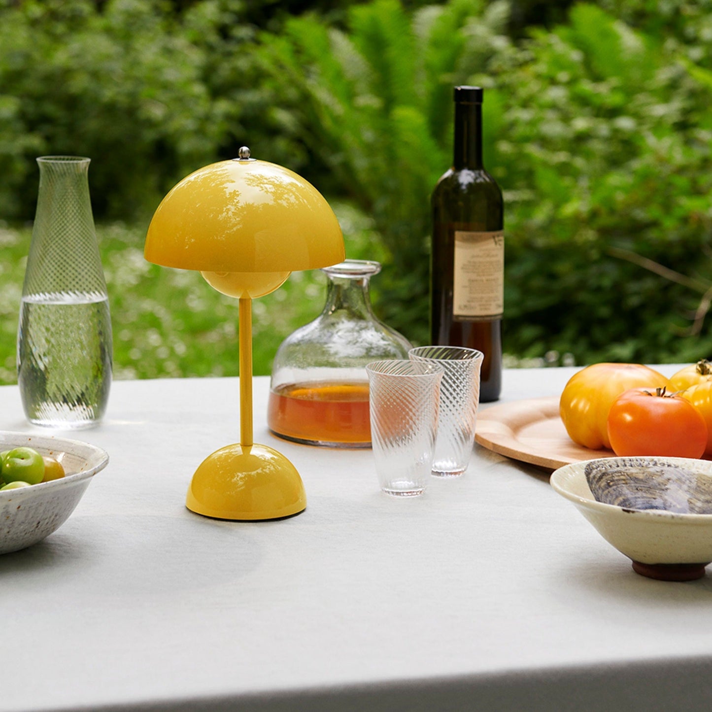 Gelbe Tischlampe auf Gartentisch mit Gläsern, Karaffe, Flasche und frischen Tomaten.