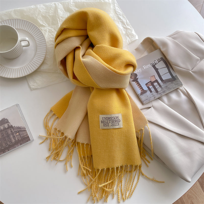 Gelber und beiger Wollschal mit Fransen auf weißem Tisch, neben Tasse und Zeitschrift.