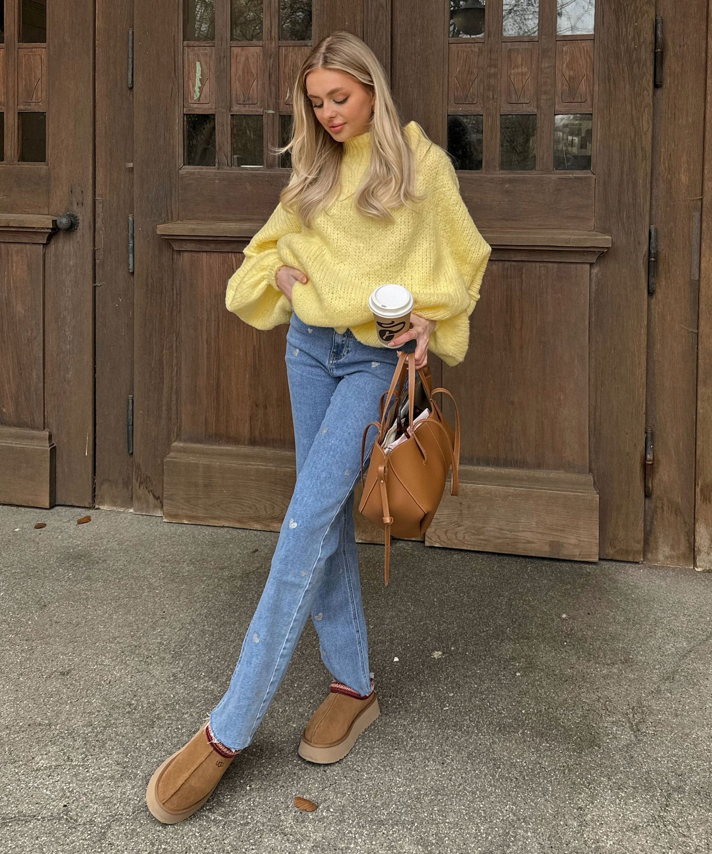 Frau in gelbem Strickpullover, blauer Jeans, braunen Schuhen, hält Kaffeebecher und Handtasche.
